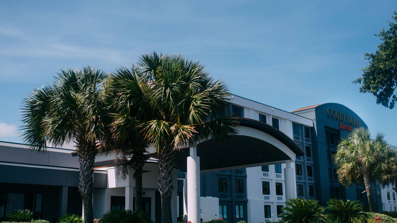 The entrance to the Courtyard by Marriott Gulfport Beachfront hotel. Several palm trees frame the awning over the hotel's main entrance.