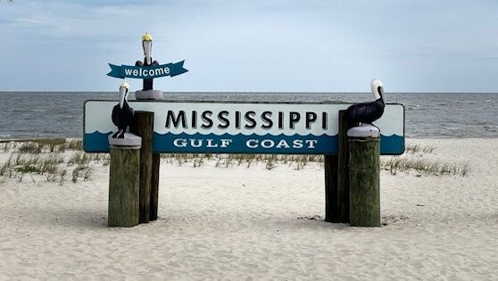 A sign reading "Welcome Mississippi Gulf Coast" on the beach with the ocean in the background. The sign is flanked by three pelican statues, one of which is holding the "Welcome" banner.
