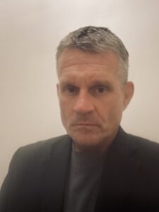 Robert Jackson's headshot. Robert is looking at the camera smiling. He has salt and pepper hair and is wearing a white shirt with a blue patterned tie and a black pinstriped suit jacket.